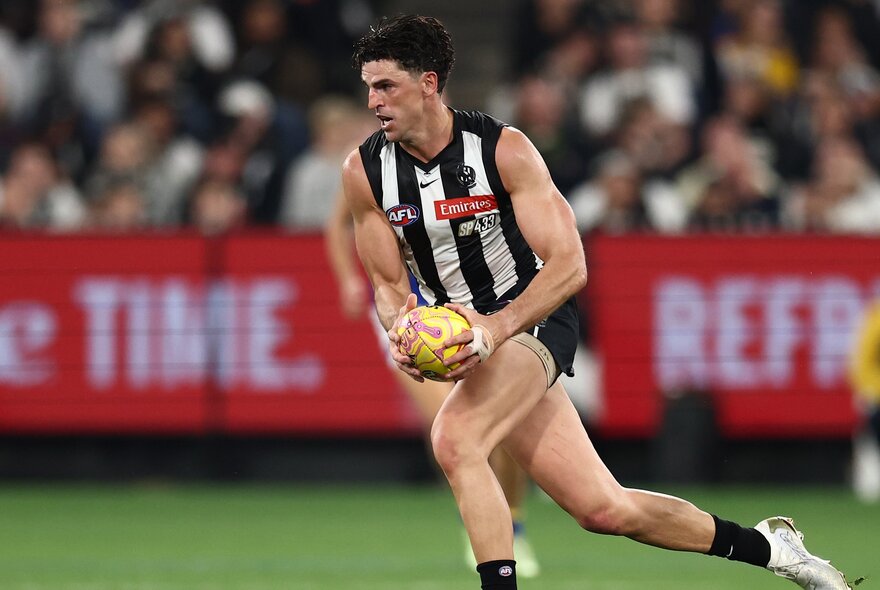 A Collingwood AFL football player running while holding the ball during a match, a blurred stadium crowd in the background.