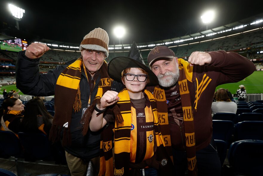 A trio of Hawthorn fans in the stadium, wearing Hawthorn team scarves and cheering.