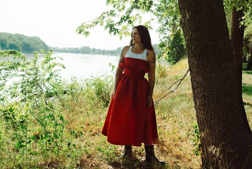 Singer Katie Crutchfield (aka Waxahatchee) wearing a red dress and standing outdoors  under a shady tree near a wide river.