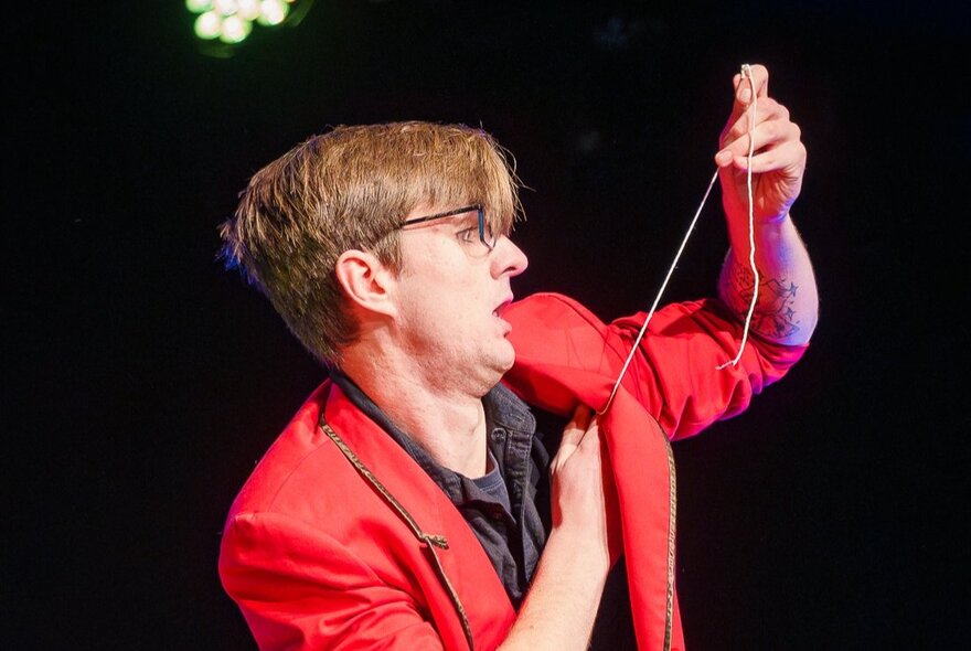 A magician performing on stage and pulling a string from underneath his red jacket. 