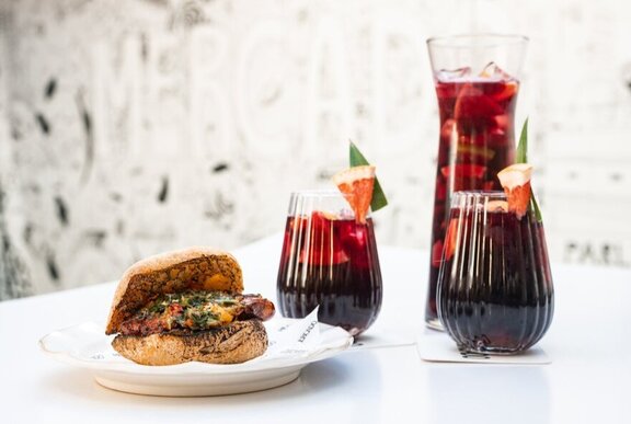 Two glasses and a carafe of sangria with a chorizo sandwich on a white plate.