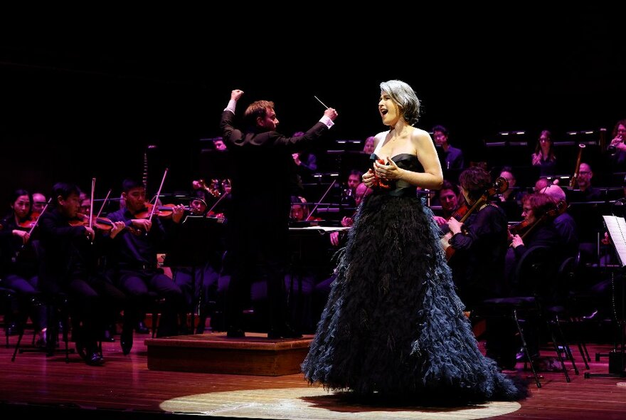 An opera singer on stage wearing an elaborate black strapless gown, with a full opera orchestra and conductor behind her.