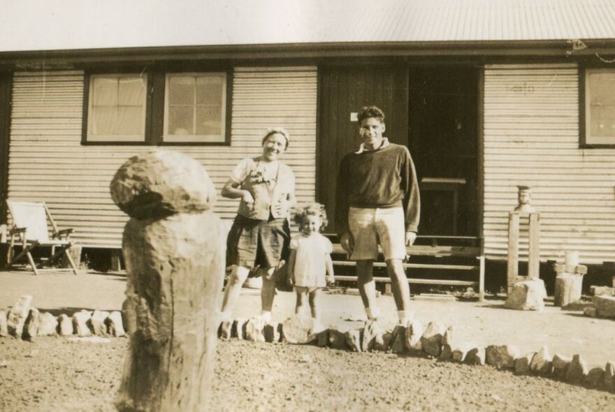 An old sepia toned image of a couple and their small child standing outside a basic wooden home.