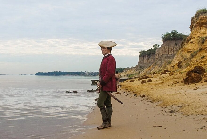 A still from a film of a person standing in profile on the shore at a beach, with sandy cliffs behind them.