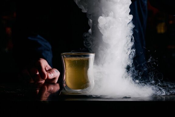 A short yellow-coloured cocktail with dry ice smoke in a column alongside it.