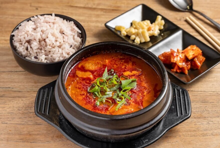 Korean kimchi soup, served in a black earthenware pot, with brown rice and condiments in separate dishes. 