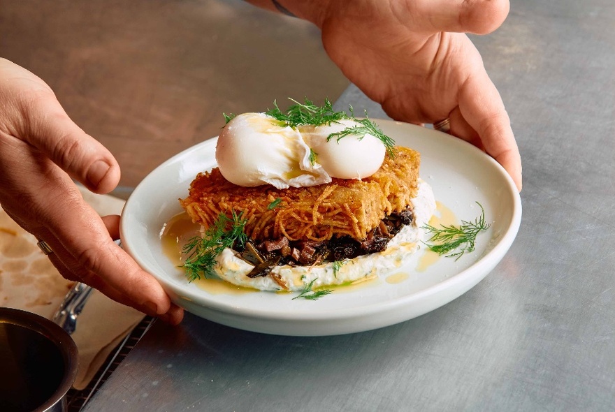 Hands putting down a plate of poached eggs and noodles on a table.