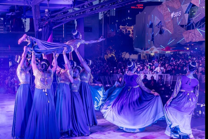 A group of performers in flowing robes dancing on the main stage at Fed Square, some of them holding a person aloft in their arms, while an audience watches from the plaza.