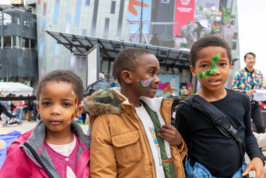 Three young children standing outdoors in the main square of Fed Square with painted faces, with people in the background.