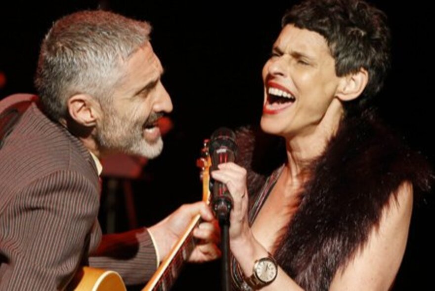 Musicians Deborah Conway and Willy Zygier singing vigorously into one microphone. 