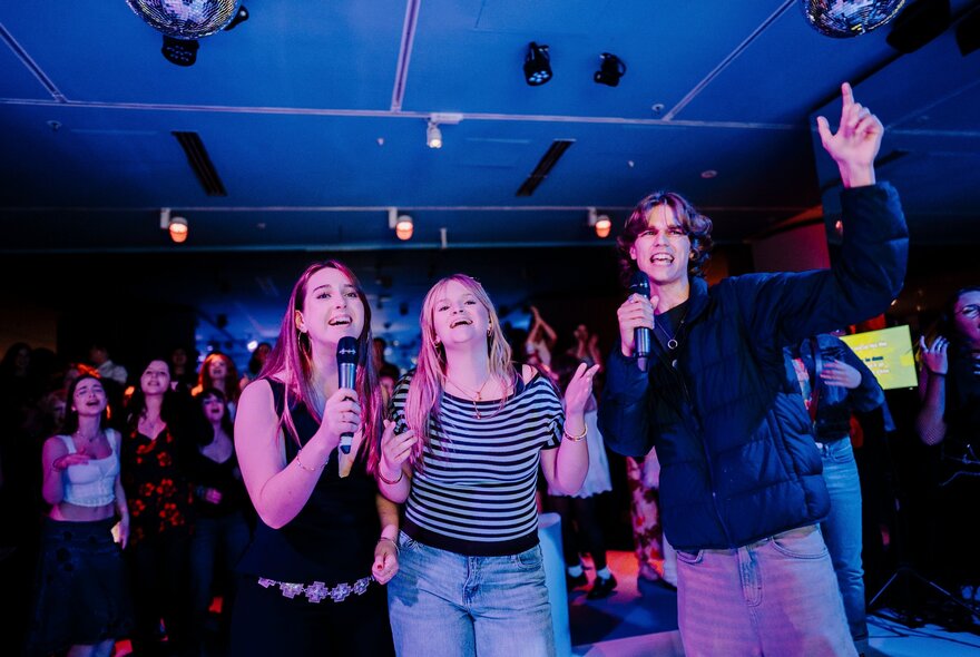 Young people holding karaoke microphones, singing and waving their hands in a crowded venue.