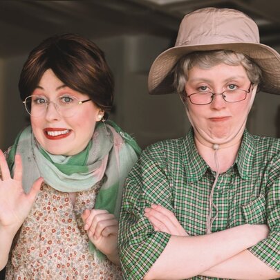 Comedians in character outfits of an older man and woman, the male wearing a hat, the woman waving her hand.