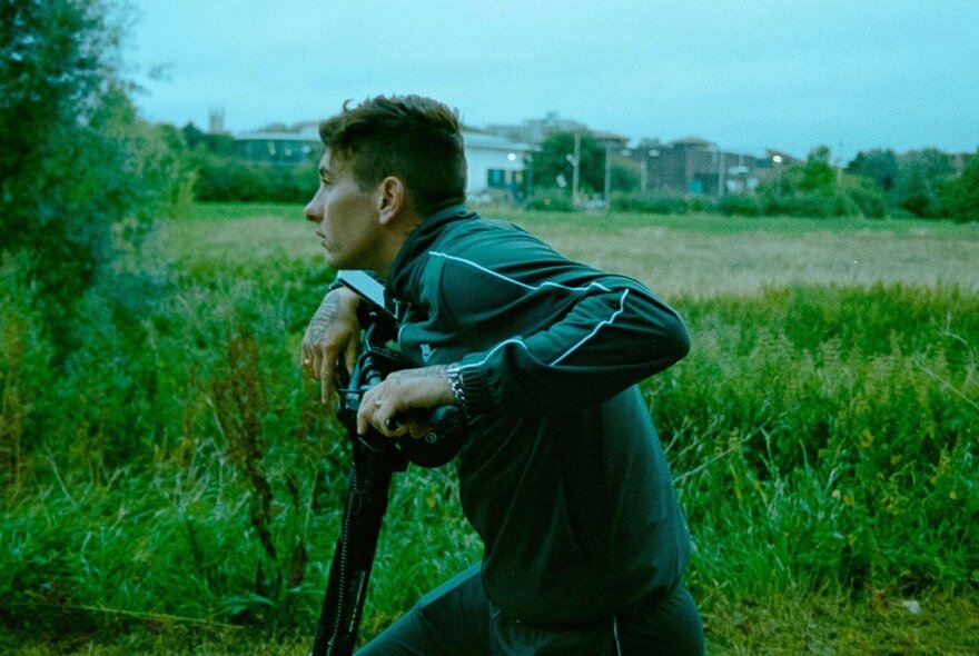 A still from a movie showing a young man with his hands resting on a scooter's handlebars, in a green field.