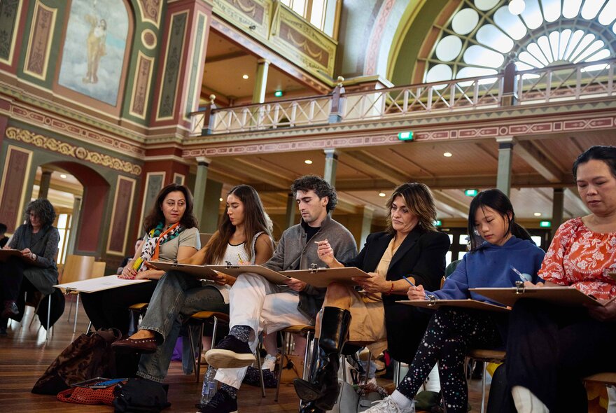 A group of people sit on chairs in rows within the ornate, high-ceilinged interior of the Royal Exhibition Building taking part in a drawing workshop.