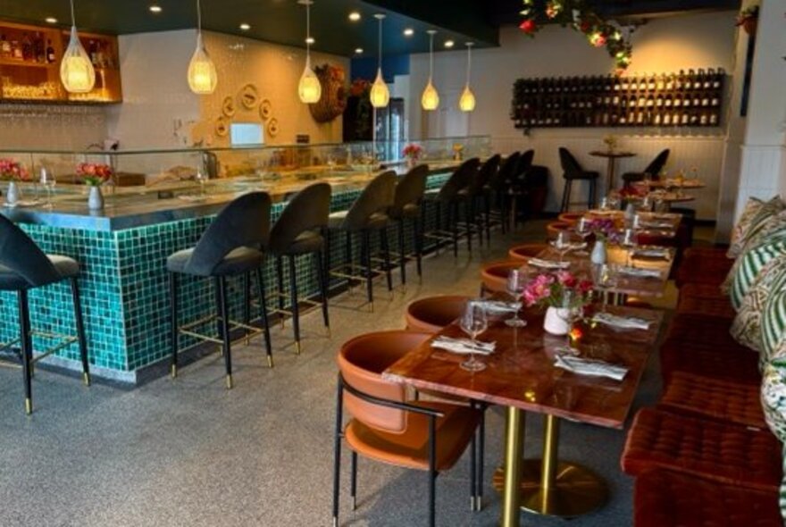 The interior of a restaurant with a row of wooden tables with curved chairs and bench seats, alongside a blue tiled bar with velvet bar stools.