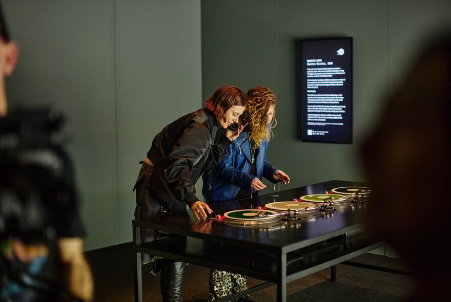 Two people leaning over a table of four turntables in an exhibition space at ACMI.