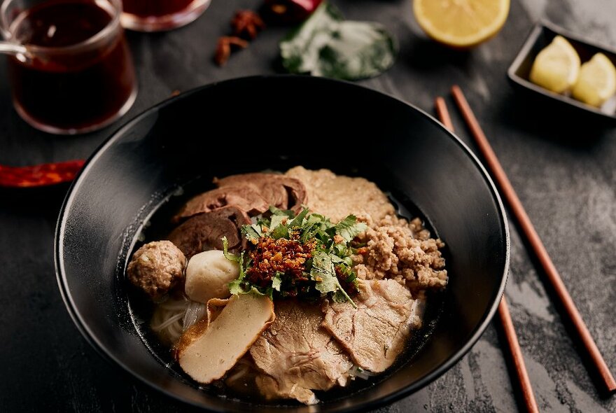 Black dish of Cambodian meat soup with chopsticks and bowls of condiments.