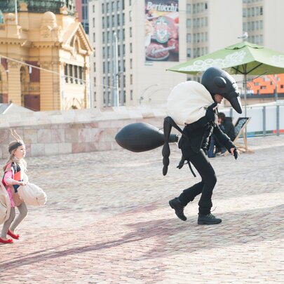 Kids following a person dressed up in an ant costume at Federation Square, Flinders Street Station visible in the background. 