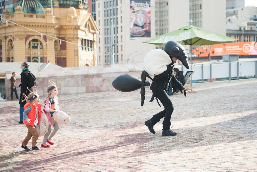 Kids following a person dressed up in an ant costume at Federation Square, Flinders Street Station visible in the background. 