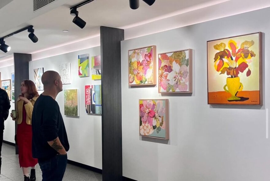 People looking at framed artworks on a white gallery wall with black lighting.