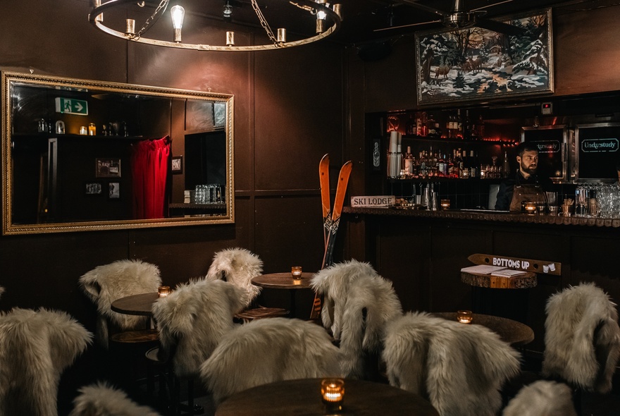 Interior of a bar with cosy Alpine decorations including fur rugs on the backs of chairs, skis resting against the bar area and candles.