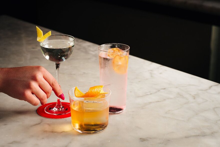 One tall-stemmed, one high-ball and one low-ball cocktail glass each with a coloured drink and fruit garnish on a marble table.