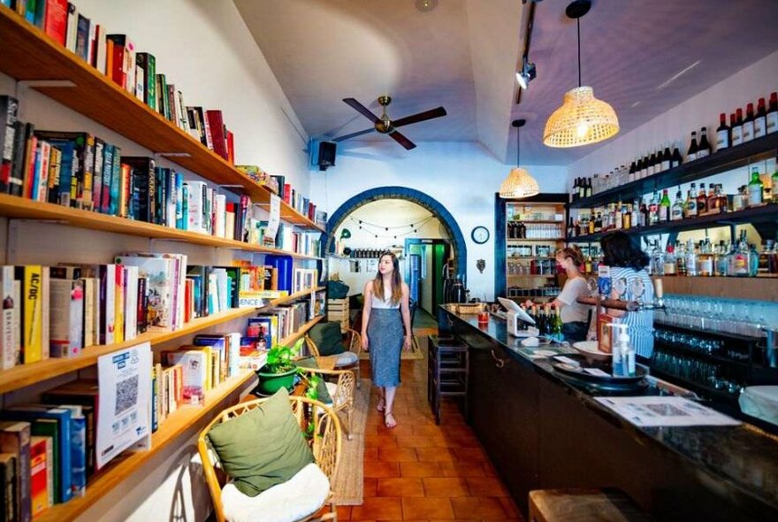 A woman walking through a wine bar with a bookshelf.