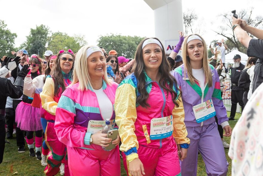 A group of charity runners smiling and wearing 1980s style sils tracksuits, walking in front of a race finish line.
