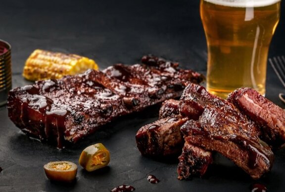 Juicy barbecue ribs next to a pint of beer on a black surface.