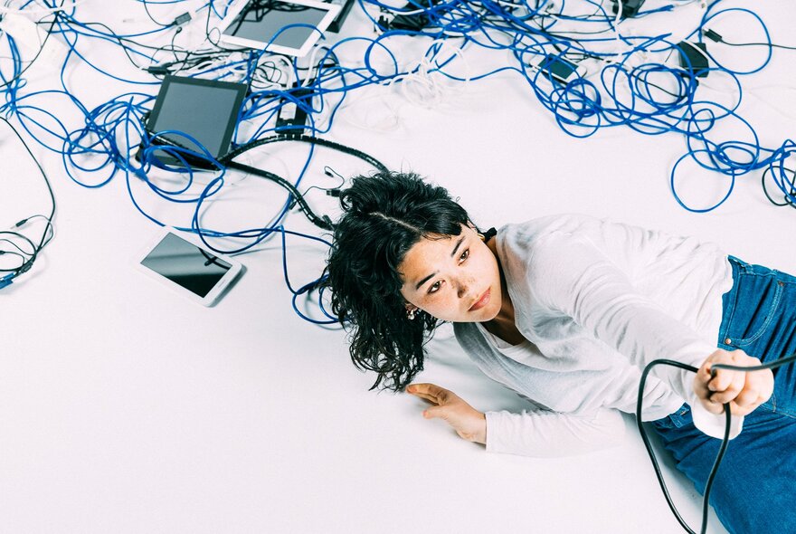 A black haired woman wearing a white shirt and blue jeans, lying on a white surface surrounded by blue, black and white cables and devices.