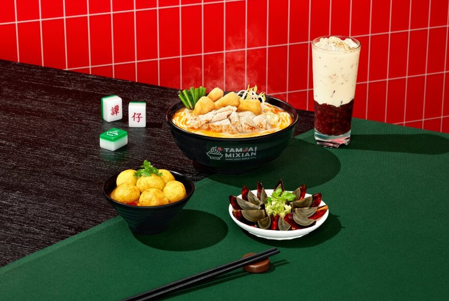 A bowl of noodle soup with sides and a drink on a green tablecloth with a red tiled wall in the backgroudn. 