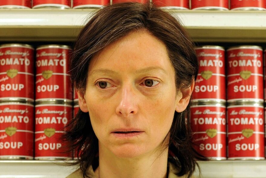 A still image from a film showing Tilda Swinton standing in front of a shelf of tins of tomato soup. 
