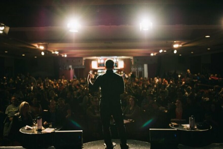 Rear view of a performer on stage at the Comic's Lounge, facing a seated audience.