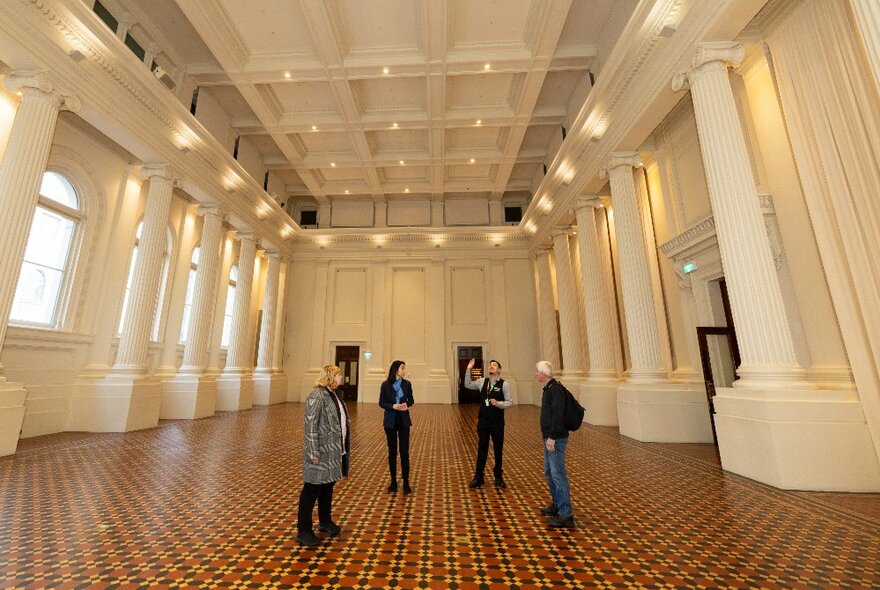 People on a small tour at the Immigration Museum, in an empty room with columns and a tiled floor.