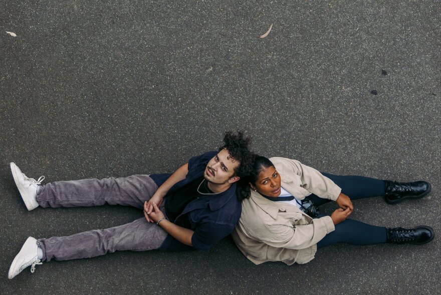 View from above of two young people sitting back to back on bitumen, their legs extended out in front of them, their faces looking up. 