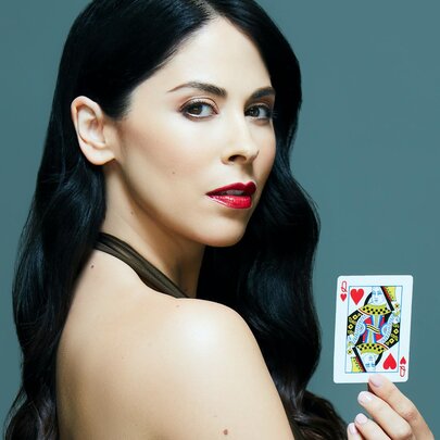 A portrait of magician Erica Vanlee with long, wavy dark hair and red lipstick, looking over her bare shoulder while holding a Queen of Hearts playing card.
