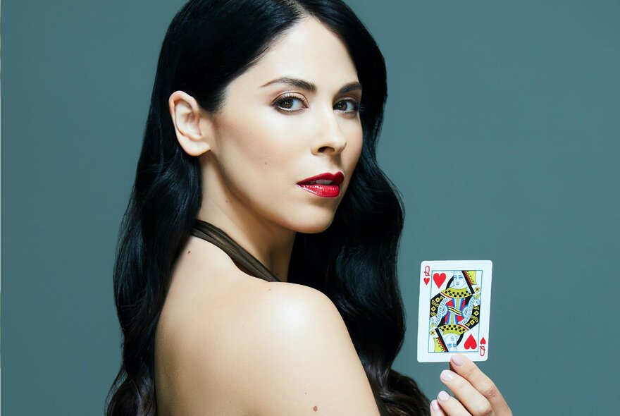 A portrait of magician Erica Vanlee with long, wavy dark hair and red lipstick, looking over her bare shoulder while holding a Queen of Hearts playing card.