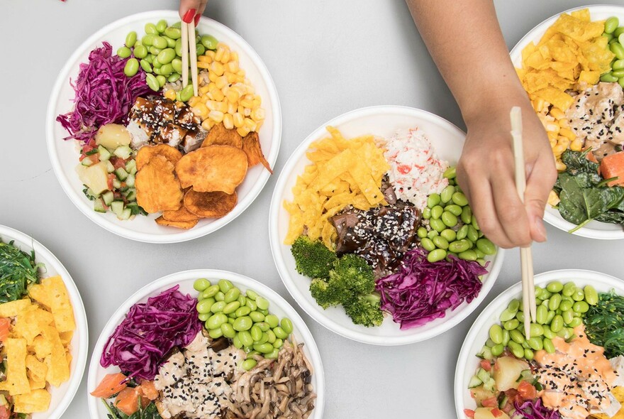 White table with plates of food, each plate with six scoops of different coloured ingredients, with hands picking up soybeans with chopsticks.