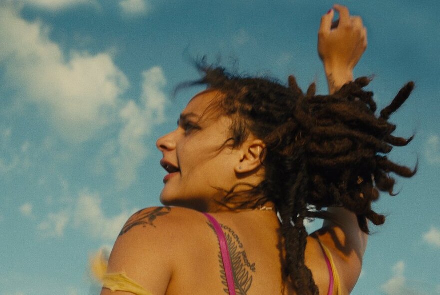 A film still of a young person outdoors with their arm raised in the air, their dreadlocked hair bouncing behind them, wearing a thin strappy top and showing a tattoo on their bare shoulder, blue sky and clouds surrounding them.