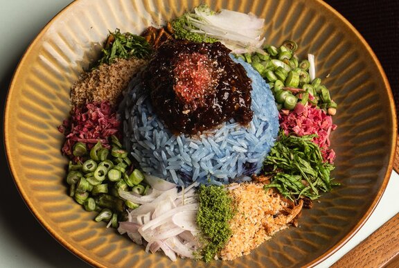 Blue rice with a selection of vegetables and garnishes in a brown dish.
