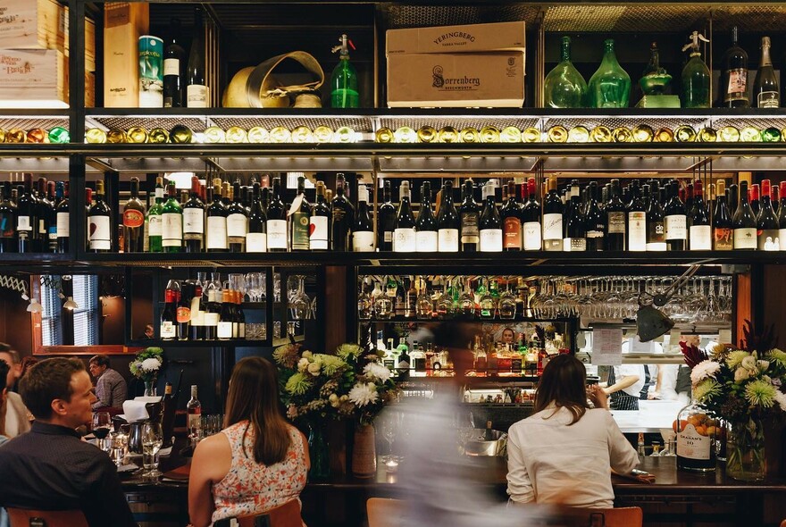 Customers lined up at a backlit bar with many shelves filled with bottles.