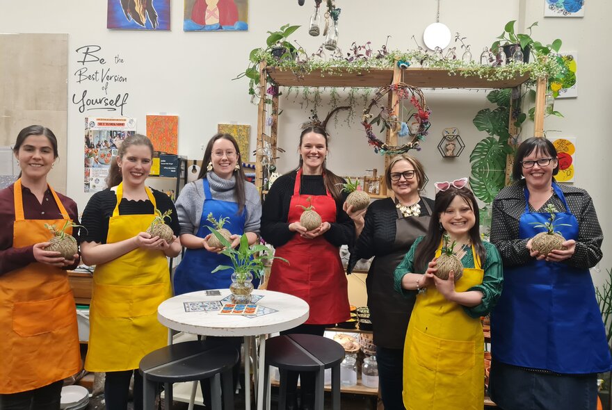 Seven people in coloured aprons holding up kokedama balls in a workshop.