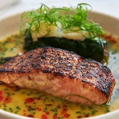 Grilled salmon alongside a dolmade with green garnish, sitting in a vegetable broth with dots of chilli oil.