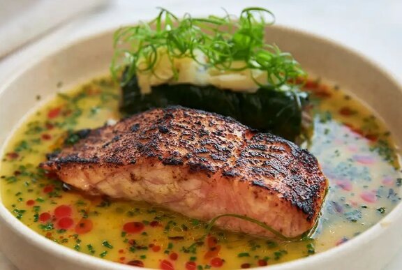 Grilled salmon alongside a dolmade with green garnish, sitting in a vegetable broth with dots of chilli oil.
