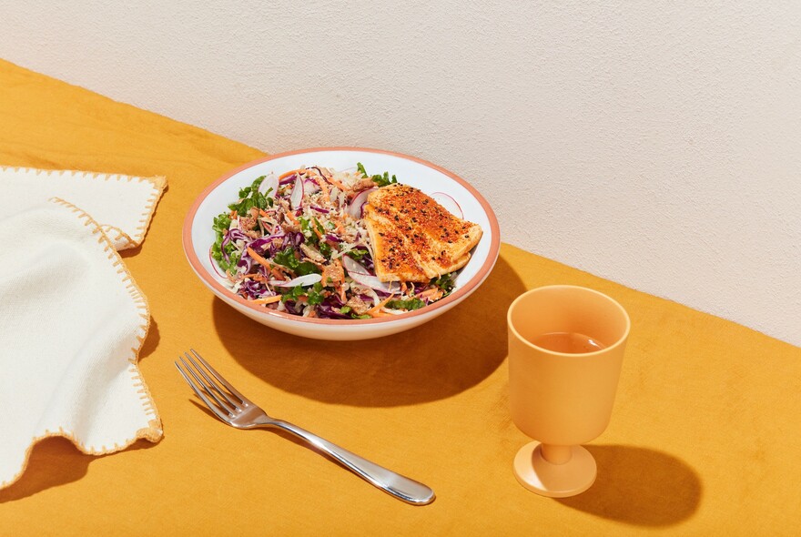 Salad bowl, serviette, fork and small yellow-orange goblet on an orange surface.