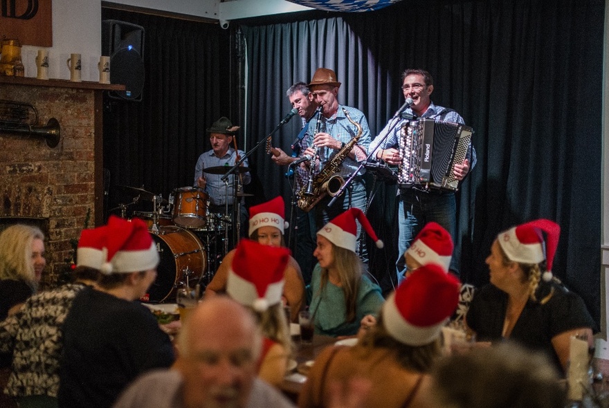 A band playing live on a small stage, in a restaurant, in front of a table of dining patrons all wearing red Santa hats. 