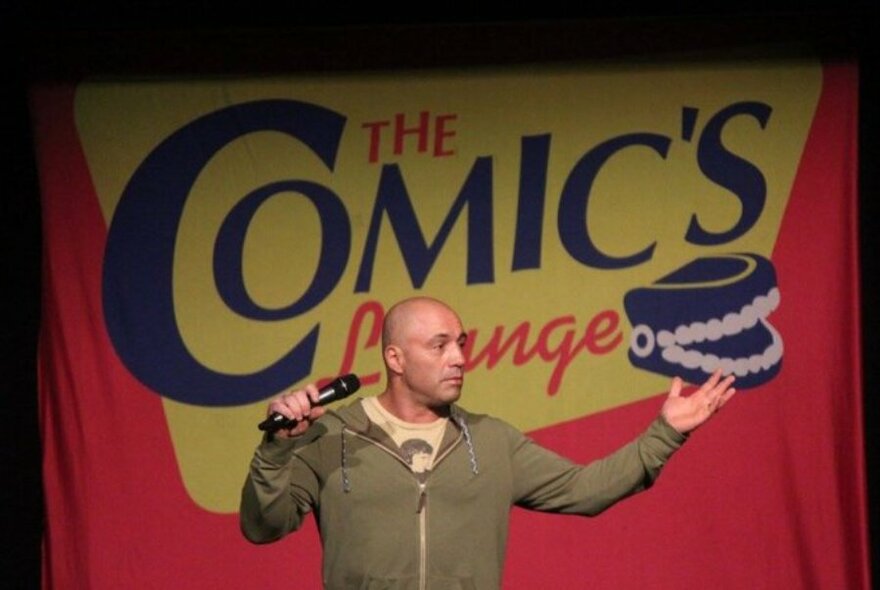 Comedian holding a microphone and performing on stage in front of a red, yellow and blue backdrop that features the words 'The Comic's Lounge' written on it.