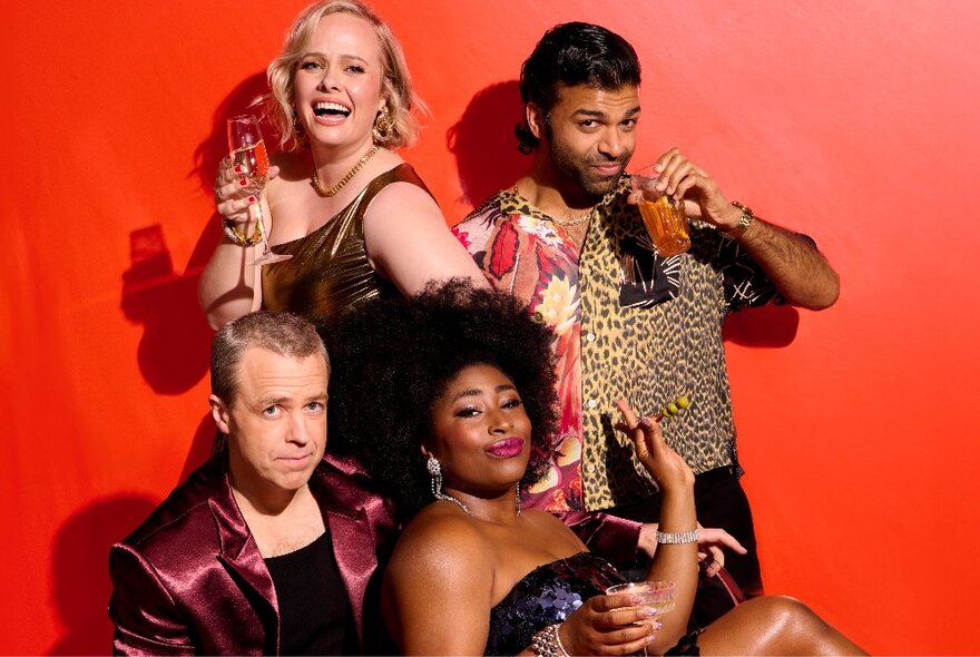 Four actors in a huddle with glasses of alcohol against a red backdrop.