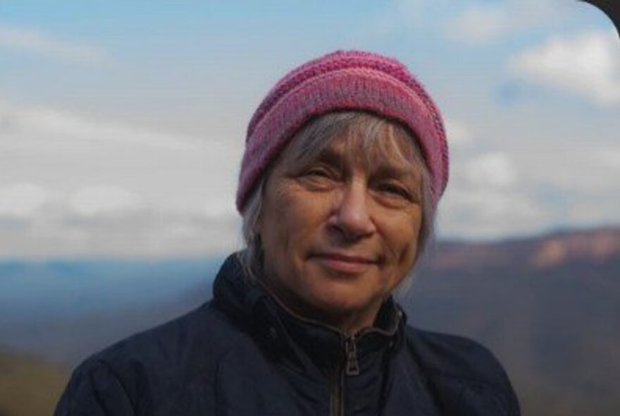 Portrait of Mununjali poet, Maria van Neerven, wearing a pink beanie and blue jacket as she smiles subtly; blurred Australian landscape as the background.