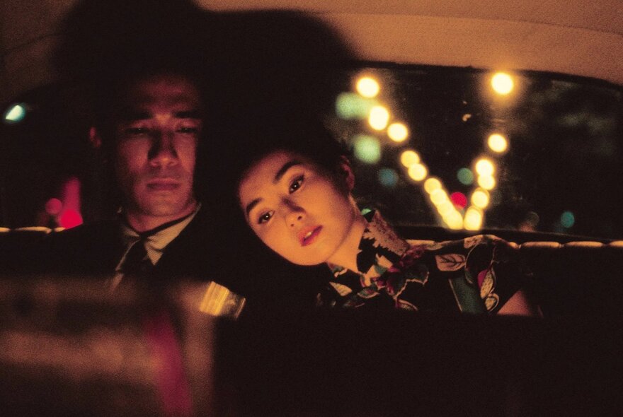A still from an Asian movie showing a couple sitting in the back seat of a car, at night.